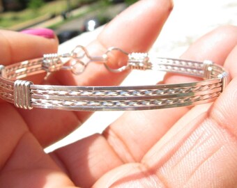 May include: A silver bracelet with a braided design. The bracelet is made of multiple strands of wire that are woven together to create a textured look.