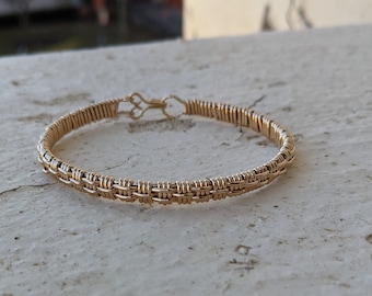 Könnte beinhalten: Ein goldfarbenes Drahtarmband mit geflochtenem Design. Das Armband besteht aus dünnem Draht, der zu einem filigranen und komplizierten Muster geflochten ist.