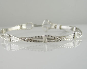 May include: A silver wire wrapped bracelet with a braided design. The bracelet is made of multiple strands of wire that are woven together to create a unique and intricate pattern.