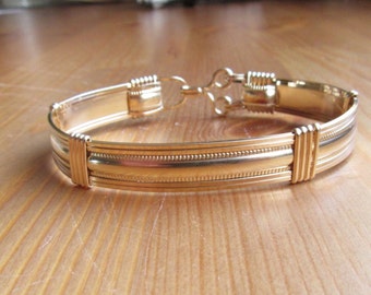 Könnte beinhalten: Ein goldenes Armband mit einem strukturierten Design und einem Verschluss. Das Armband liegt auf einer Holzoberfläche.