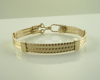 Könnte beinhalten: Ein goldfarbenes Armband mit Hakenverschluss. Das Armband hat eine Reihe paralleler, mit Perlen besetzter Bänder und gewickelte Drahtdetails. Das Design ist symmetrisch, mit Fokus auf die filigrane Metallarbeit und Handwerkskunst.