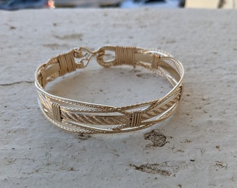 Könnte beinhalten: Ein goldfarbenes, drahtgewickeltes Armband mit geflochtenem Design. Das Armband besteht aus mehreren Drähten, die miteinander verflochten sind, um ein einzigartiges und kompliziertes Muster zu schaffen.