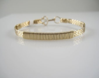 Könnte beinhalten: Ein goldfarbenes Drahtarmband mit gewebtem Muster. Das Armband hat einen einfachen Verschluss.