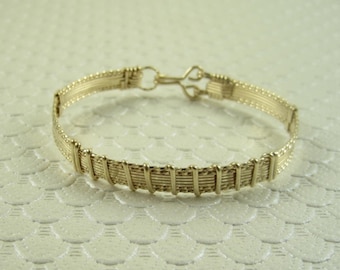 Könnte beinhalten: Ein goldfarbenes Drahtarmband mit gewebtem Muster. Das Armband hat einen einfachen Verschluss.