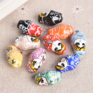 May include: Assortment of colourful ceramic beads with painted faces and designs. The beads are oval shaped and feature various colours including yellow, blue, pink, red, orange, and black.