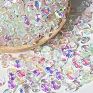 May include: A large quantity of iridescent clear glass beads in a teardrop shape. The beads are scattered on a white surface.