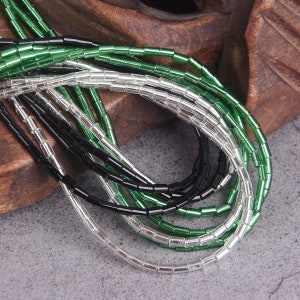 May include: A close-up of a pile of green, black, and silver glass beads. The beads are small and cylindrical, and they are arranged in a random pattern.