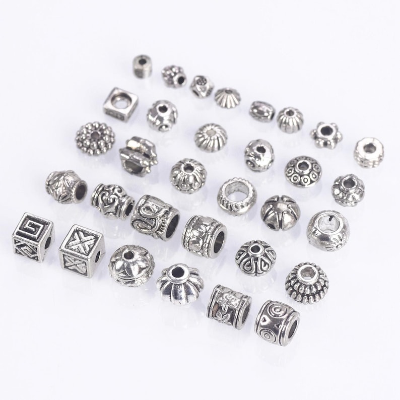May include: Assortment of silver metal beads with various designs and shapes, including round, square, and cylindrical beads.
