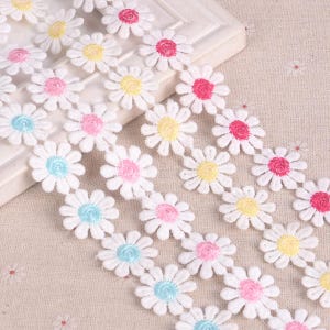 May include: White lace trim with a repeating pattern of small embroidered daisies in pink, yellow, blue, and red.