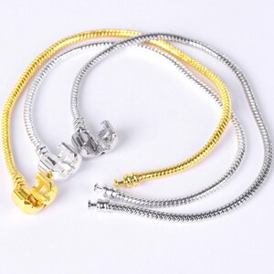 Puede incluir: Tres pulseras de cadena de serpiente en tonos dorados y plateados. Las pulseras tienen cierres. La pulsera dorada tiene un cierre en forma de corazón. La pulsera plateada tiene un cierre redondo. Las pulseras se muestran sobre un fondo blanco.