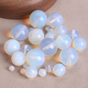 May include: A selection of opalescent, round beads in various sizes. The beads are mainly white with delicate blue and white stripes, and have a glossy finish. Some beads have a small hole for stringing. The beads are scattered on a neutral surface.