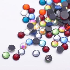 May include: A collection of small, round, multi-colored flat-back rhinestones in shades of red, blue, green, orange, and clear. Each piece features a faceted top surface and a dark, textured, circular base, scattered across a plain white surface.