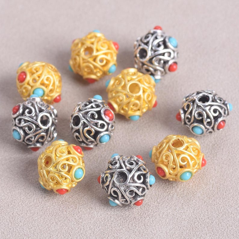 May include: A collection of filigree beads in gold and silver tones. Each bead features an openwork design with red and turquoise accents. The beads are spherical and appear to be made of metal.