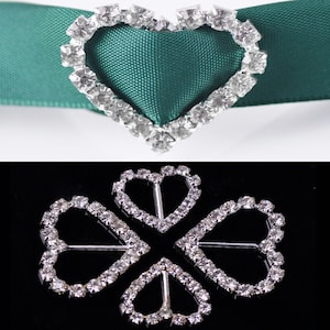 Puede incluir: Dos adornos de corazón de plata con pedrería. Uno es un corazón único, el otro es un diseño de trébol de cuatro hojas con cuatro corazones conectados.