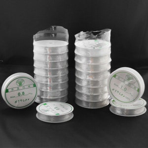 May include: Two stacks of clear elastic string spools, each spool labelled with the text "Size 0.8" and "Size 1.0" in Japanese. The spools are packaged in clear plastic.