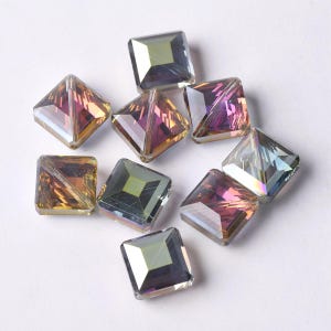 Puede incluir: Un conjunto de ocho cuentas de vidrio de forma cuadrada con una superficie facetada. Cada cuenta muestra un acabado iridiscente con tonos cambiantes de púrpura, oro y plata, creando una apariencia reflectante en las caras geométricas.