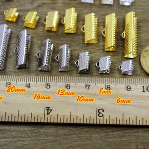 May include: Assortment of gold and silver metal clasps in various sizes, measured in millimeters, for jewelry making. The clasps are arranged on a wooden ruler.