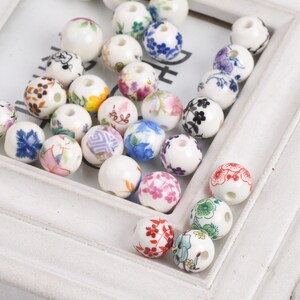 May include: A collection of white ceramic beads with various floral patterns in shades of blue, pink, red, yellow, and green. The beads have a hole in the center for stringing.