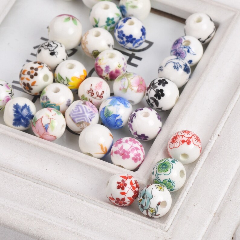 May include: A collection of white ceramic beads with various floral patterns in shades of blue, pink, red, yellow, and green. The beads have a hole in the center for stringing.