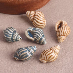 Peut inclure: Un ensemble de six perles en céramique en forme de coquillages en spirale. Trois perles présentent une finition brillante rayée beige et marron, tandis que les trois autres affichent un motif rayé bleu et gris brillant. Chaque perle est percée.