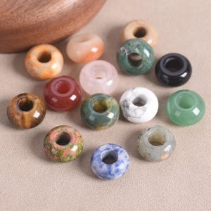 May include: A collection of fourteen smooth, round, donut-shaped beads made from various polished stones. The beads feature diverse colors and patterns, including shades of orange, green, white, black, brown, red, pink, and blue, each with a central hole.