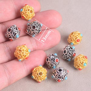 May include: A collection of decorative beads, each approximately 0.5 inches in diameter. The beads feature intricate openwork designs in gold and silver tones, accented with red and turquoise-colored stones. The beads are displayed against a neutral background.