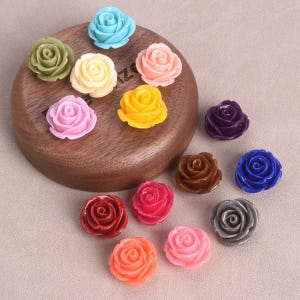 Puede incluir: Surtido de 12 adornos de flores de rosa de resina de colores. Las flores son de varios colores, incluyendo rosa, azul, amarillo, verde, morado, marrón y rojo. Las flores son todas del mismo tamaño y forma, y están dispuestas en círculo sobre una superficie de madera.