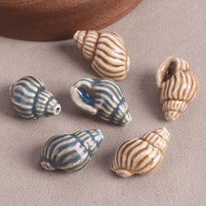May include: Six ceramic seashell beads in shades of blue, white, and tan. The beads feature detailed spiral patterns and vary in size and shape. These decorative shells are suitable for jewellery making or coastal home decor.