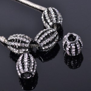 May include: A set of four silver charm beads with a black metal base and clear rhinestones. The beads are oval shaped and have a striped design.