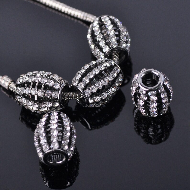 May include: A set of four silver charm beads with a black metal base and clear rhinestones. The beads are oval shaped and have a striped design.