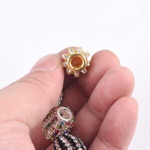 May include: A gold-toned bead with a clear crystal design and a round hole in the center. The bead is being held in a hand.