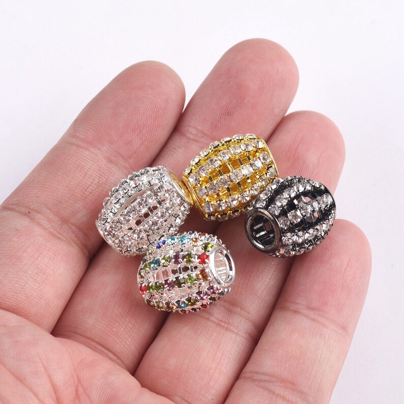 May include: Four different colored metal barrel-shaped beads with clear and colored rhinestones. The beads are in a hand.