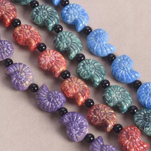 Peut inclure: Gros plan de plusieurs brins de perles de pierres précieuses. Les perles sont sculptées en forme de coquillage en spirale et sont de différentes couleurs, notamment rouge, bleu, vert et violet. Des perles noires séparent les perles en forme de coquillage.