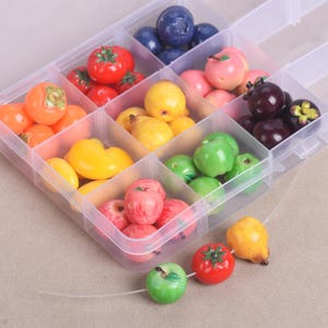 Könnte beinhalten: Ein durchsichtiger, unterteilter Kunststoff-Aufbewahrungsbehälter gefüllt mit verschiedenen Miniatur-Obstperlen, darunter rote Tomaten, gelbe Birnen, grüne Äpfel, blaue Blaubeeren und dunkelviolette Mangostane, mit drei auf einen Faden aufgezogenen Perlen.