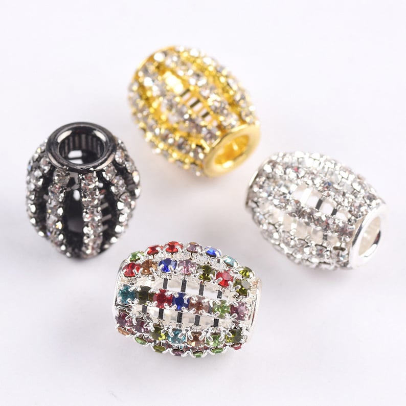 May include: Four different colored metal barrel-shaped beads with a cage design and clear and colored rhinestones. The beads are on a white surface.