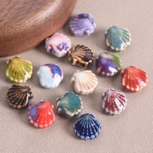 Pode incluir: Uma coleção de contas de cerâmica em forma de concha em várias cores, incluindo azul, vermelho, branco e castanho. As contas têm um acabamento brilhante e estão dispostas numa superfície neutra. Ideais para a criação de joias e projetos de artesanato.
