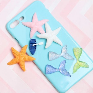 May include: A blue phone case decorated with various sea-themed charms, including pink, white, and orange starfish, blue and green mermaid tails, and a small blue boat.