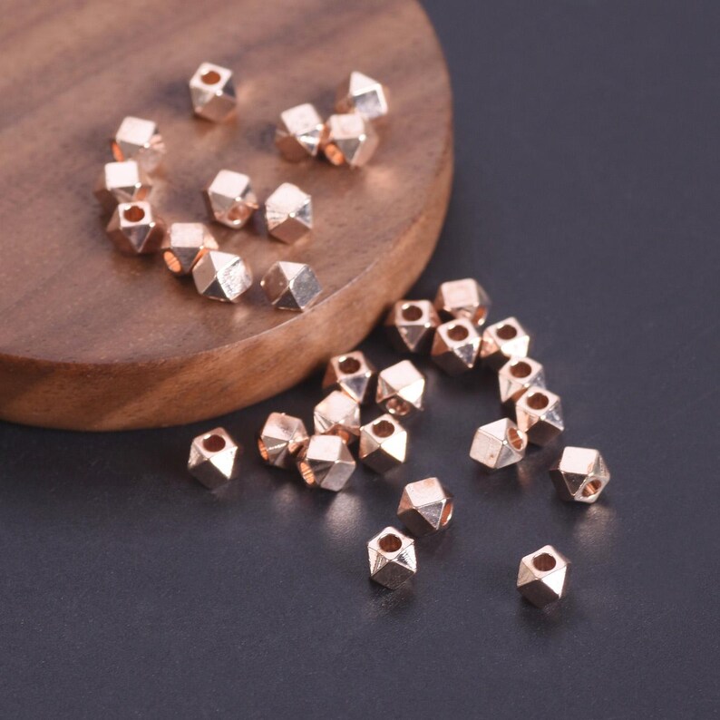 Pu&ograve; includere: Una collezione di piccole perline color oro rosa con una forma geometrica sfaccettata. Ogni perlina presenta un foro centrale per l'infilatura. Le perline sono sparse su una superficie scura, alcune appoggiate su una base circolare in legno.