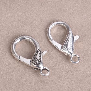 Könnte beinhalten: Zwei silberfarbene Metall-Hummerverschlüsse mit verzierten Designs. Die Verschlüsse sind offen und bereit, an einer Kette oder anderem Schmuck befestigt zu werden.