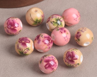 May include: A collection of round beads in shades of pink and cream, adorned with floral designs in deep pink and green. The beads vary in size and have a glossy finish, perfect for jewelry making.