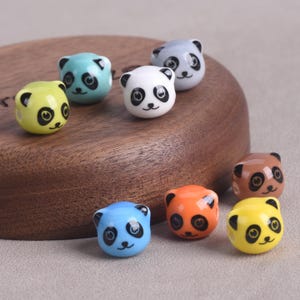 Puede incluir: Una colección de cuentas con forma de panda de colores, incluyendo blanco, azul, amarillo, naranja y marrón. Cada cuenta presenta una cara de panda en blanco y negro. Las cuentas están dispuestas sobre una superficie de madera.