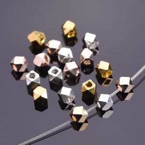 Pu&ograve; includere: Una collezione di piccole perline geometriche a pi&ugrave; facce con finiture metalliche color oro, argento e oro rosa. Diverse perline sono sparse, mentre tre sono infilate su un sottile filo monofilamento trasparente.