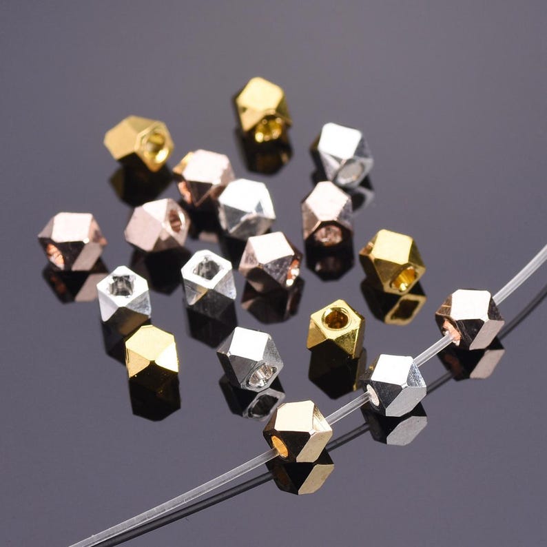Pu&ograve; includere: Una collezione di piccole perline geometriche a pi&ugrave; facce con finiture metalliche color oro, argento e oro rosa. Diverse perline sono sparse, mentre tre sono infilate su un sottile filo monofilamento trasparente.