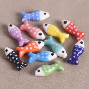May include: A collection of eleven small, fish-shaped ceramic beads in various colors including blue, pink, yellow, red, orange, light blue, black, green, and dark blue. Each bead features a white head with a black eye and a polka-dot pattern.