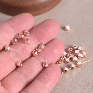 Pu&ograve; includere: Una collezione di piccole perline metalliche color oro rosa con una forma geometrica sfaccettata. Diverse perline sono sparse sulla superficie, mentre altre sono disposte in fila su un sottile filo metallico, mostrando la loro superficie riflettente.