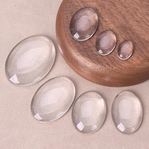 Peut inclure: Cinq cabochons ovales en verre transparent de tailles différentes, présentés sur une surface en bois. Les cabochons sont lisses et translucides, avec un léger chatoiement.