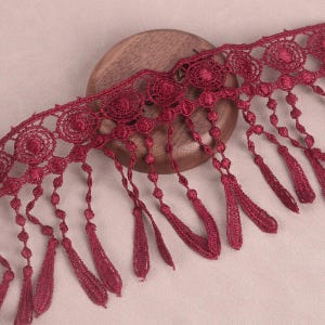 May include: Burgundy red lace trim with a scalloped edge and fringe tassels. The trim has a delicate floral pattern.