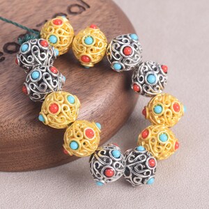 May include: A collection of decorative beads in gold and silver tones, adorned with red and turquoise accents. The beads feature intricate openwork designs and are arranged in a circular pattern, showcasing their detailed craftsmanship.