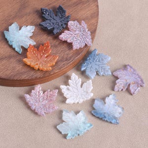 May include: A collection of colorful maple leaf-shaped embellishments. The leaves are in various shades, including blue, orange, pink, and white, with glitter accents. These decorative pieces are ideal for crafting and jewelry making.