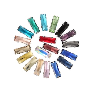 May include: Assortment of colourful rectangular glass rhinestones with silver settings. The rhinestones are arranged in a circle on a white background.